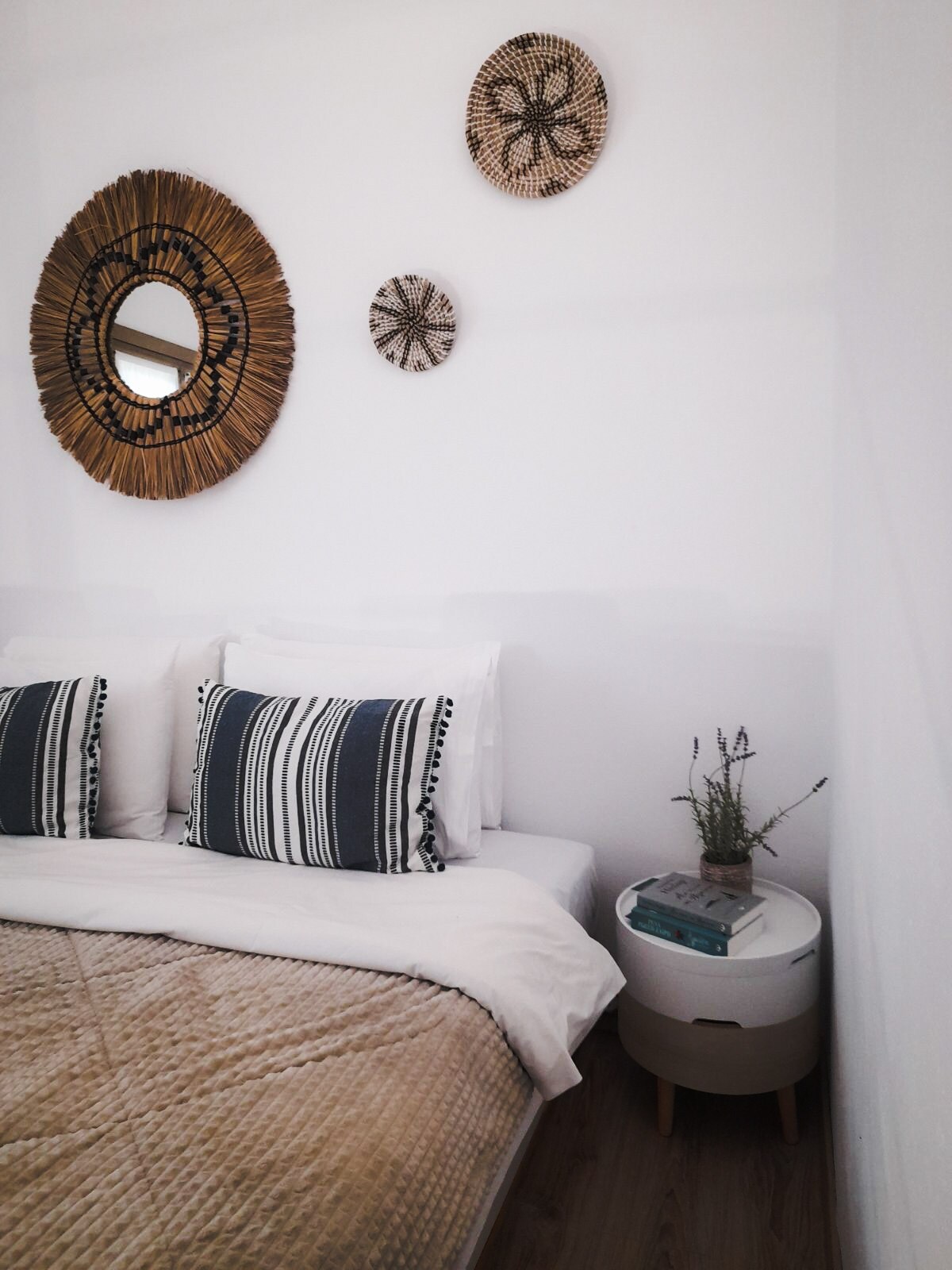 A sleeping area features a spacious bed dressed in neutral linens, complemented by decorative pillows. Above the bed, woven wall decor adds texture to the bright white walls. A small bedside table holds a book and a plant, enhancing the serene environment.