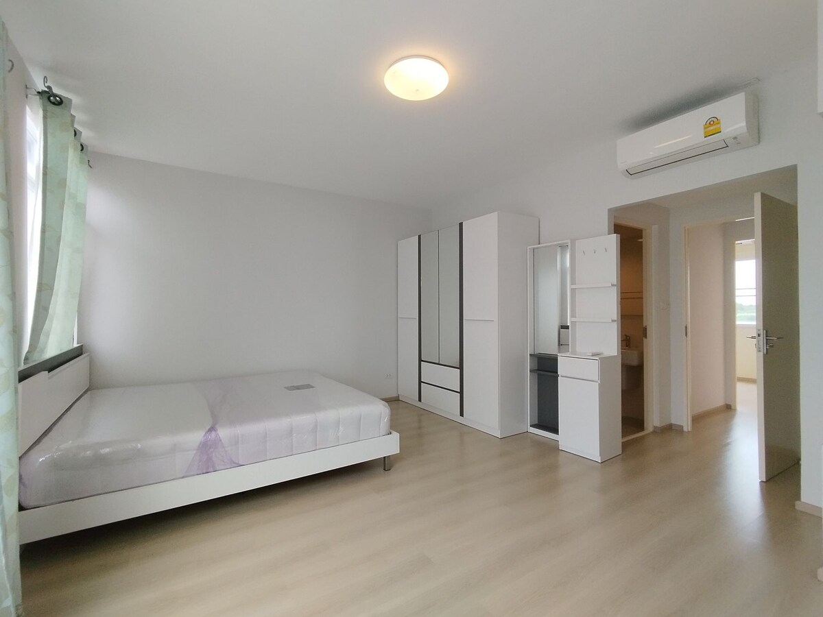 A spacious room is presented with light-colored walls and wooden flooring. A large bed is positioned against one wall, while a wardrobe and a small shelving unit are visible. Natural light enters through a window with sheer curtains, enhancing the open atmosphere.