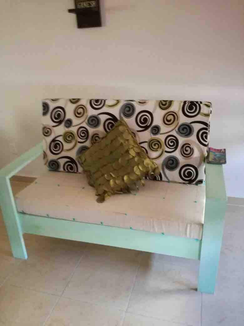A light green chair is positioned against a neutral wall. The seat features a padded cushion, topped with a decorative pillow in a textured design. The backrest displays a fabric with a swirling pattern in various colors, contributing a playful touch to the seating area.