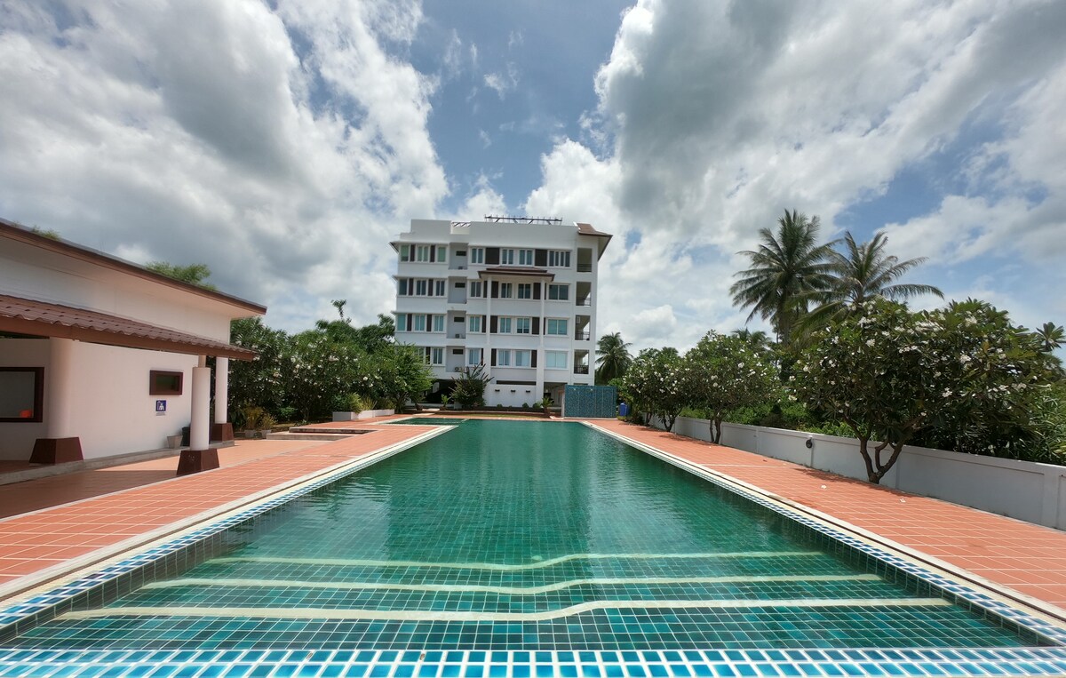 A spacious pool is framed by patterned tiles and surrounded by greenery, with a multi-story building rising in the background. The sky features large clouds, adding dimension to the serene setting.