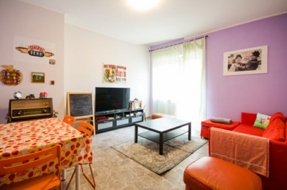 A vibrant living area is presented, featuring a red sofa, a coffee table, and a small dining table with orange chairs. A television is positioned on a low media unit under a large window, providing natural light that fills the space.