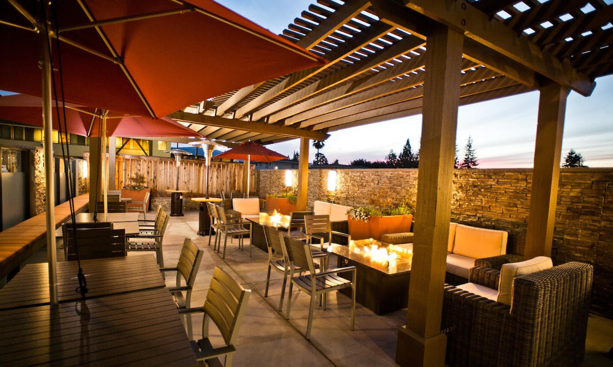 An outdoor patio space features a mix of seating arrangements, including lounge chairs and small dining tables, shaded by red umbrellas. A stone wall creates a cozy backdrop, while flickering flames from fire pits add warmth to the inviting area, set against a twilight sky.