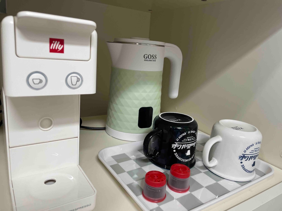 A compact kitchen area features an Illy coffee machine alongside a green electric kettle. Two mugs are positioned on a tray, accompanied by coffee capsules, providing essentials for a comfortable beverage experience.