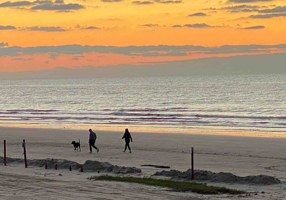 Two individuals are seen walking along the beach at sunset, accompanied by a dog. Soft colors of orange and pink fill the sky, reflecting on the gentle waves of the Gulf. The sandy shoreline is accentuated by footprints, indicating a serene stroll in the tranquil atmosphere.