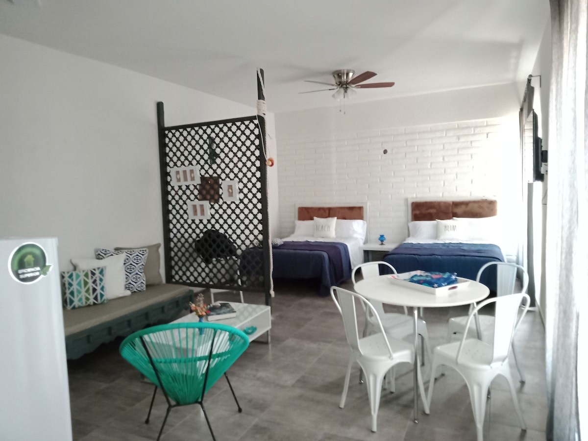A modern interior space features two spacious beds with wooden headboards and blue bedding. A lounge area includes a green chair and a couch facing a decorative wall. A round dining table and chairs are positioned near a light-filled window, creating a functional living area.