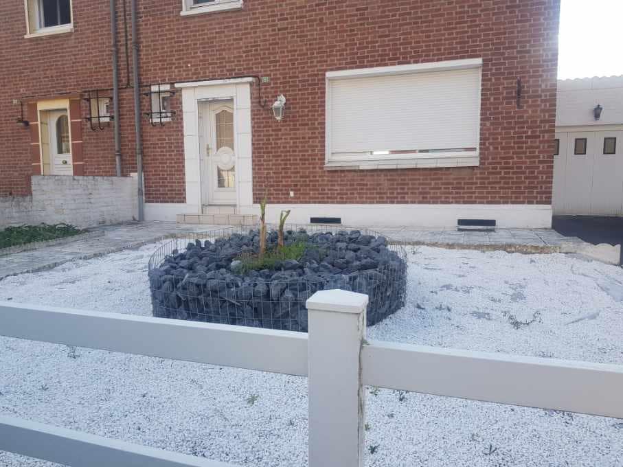 The exterior features a low-maintenance yard with gravel and a circular rock formation surrounding a small tree. A brick facade is visible on the house, with a large window partially obscured by a roller shutter. The front entrance is marked by a door with a unique design.