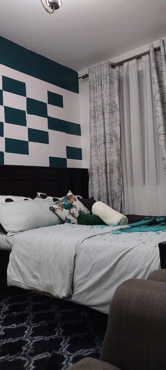 A cozy bedroom features a dark wooden headboard supporting a neatly made bed with soft pillows. The wall behind displays an abstract pattern of teal and white squares. Light gray curtains frame a window, allowing natural light to fill the room.