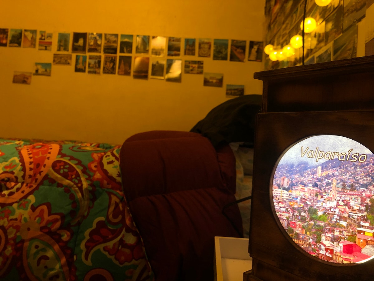 A vibrant sleeping area is highlighted by a colorful patterned blanket draped over the bed. A bedside table features a vintage-style lamp displaying a scenic view of Valparaíso. Behind the bed, a collection of postcards adorns the wall, enhancing the room's character.