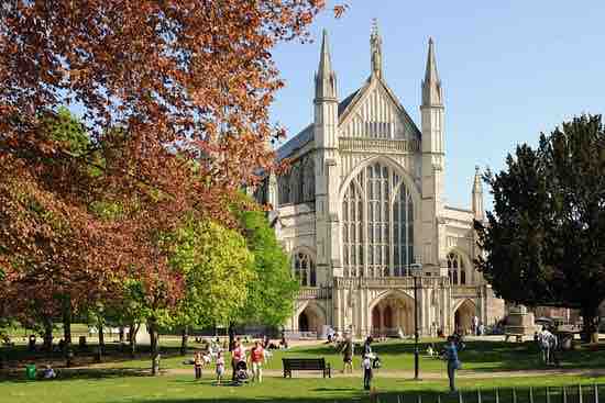 A grand cathedral with intricate Gothic architecture rises against a clear blue sky. Surrounding greenery features blooming trees and manicured lawns, where visitors enjoy leisure activities. Benches provide seating options for those wishing to relax and take in the serene atmosphere of the park.