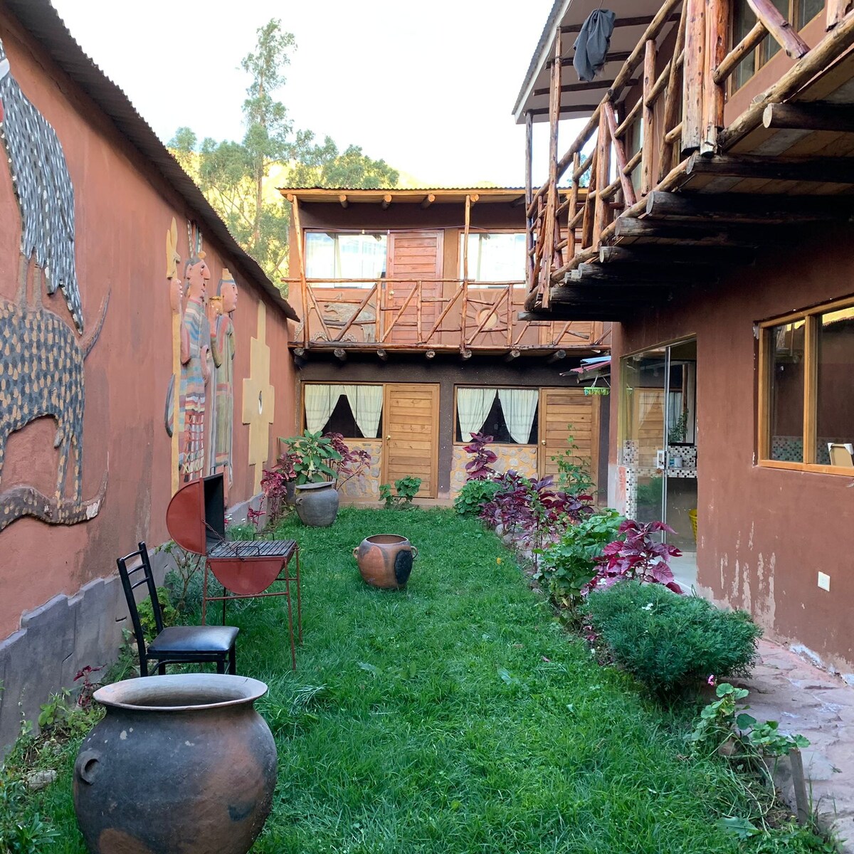 A serene courtyard is surrounded by colorful murals and natural stone walls. The grassy area features a blend of plants and decorative pots. Wooden balconies extend above, while a few simple chairs and tables provide seating near the vibrant green space.