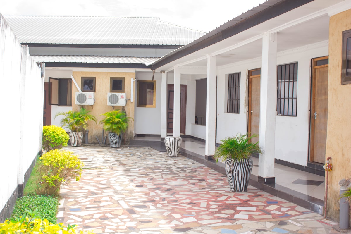 A welcoming courtyard features a mosaic stone pathway surrounded by well-maintained greenery. Guests can find air conditioning units mounted on the exterior walls, along with several doors leading into the self-contained rooms. Potted plants are positioned strategically for added touch.