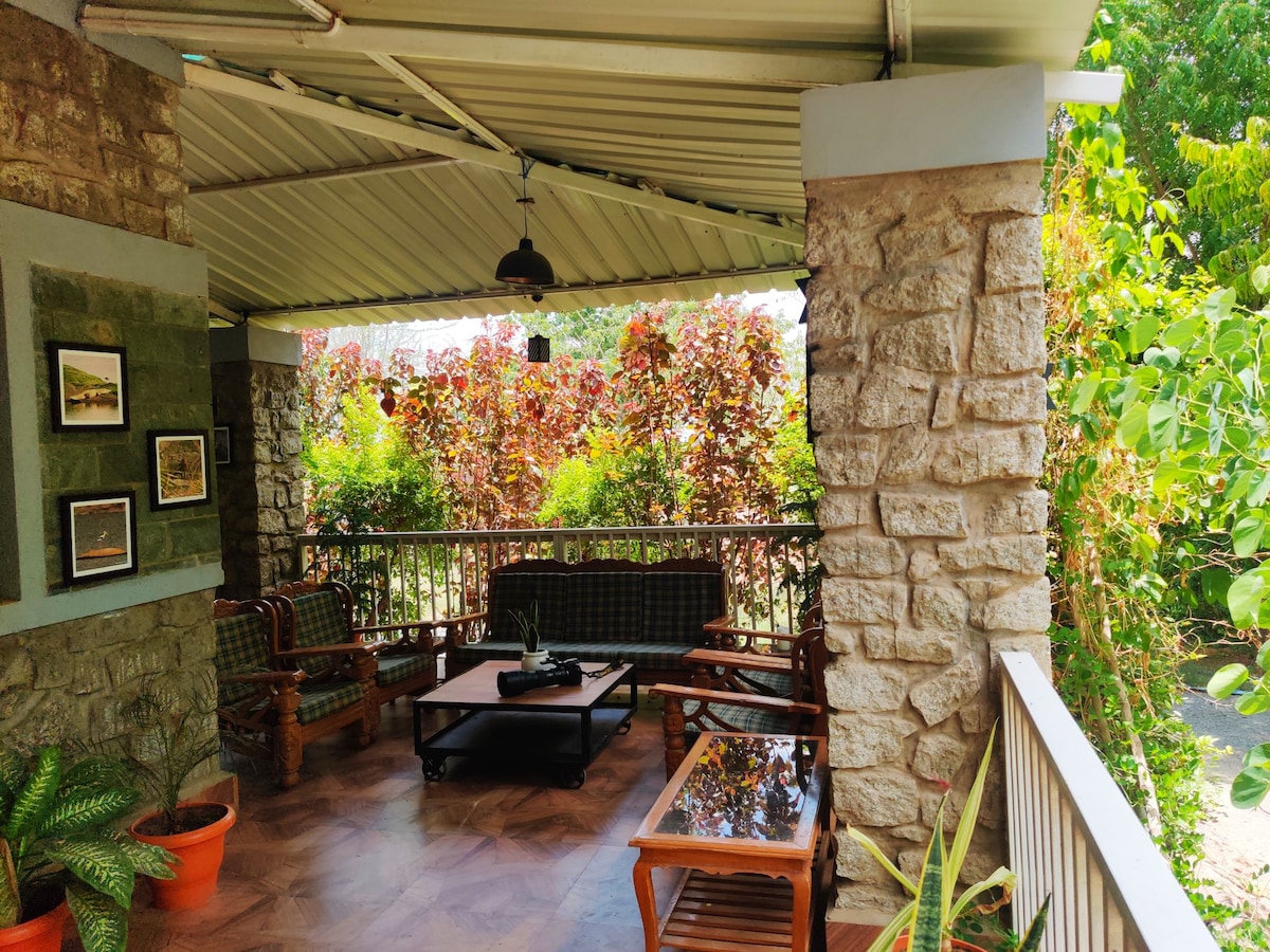 An inviting outdoor veranda features wooden furniture, including a coffee table and comfortable seating. Lush greenery surrounds the space, with vibrant plants creating a peaceful atmosphere. Natural light illuminates the area, enhancing the stone walls and tiled floor.
