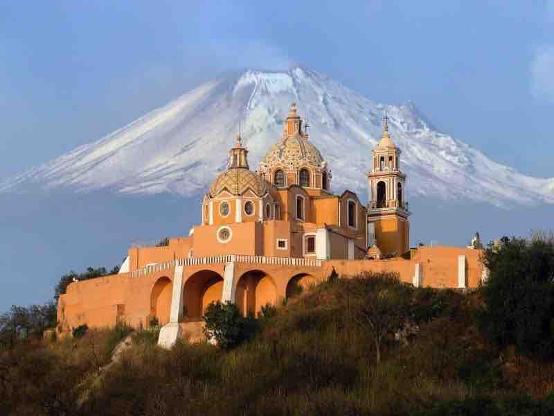 A historic building with distinct domes and bell towers is situated on an elevated landscape. In the background, the snow-capped peak of a mountain is prominently visible, contrasting against the blue sky.