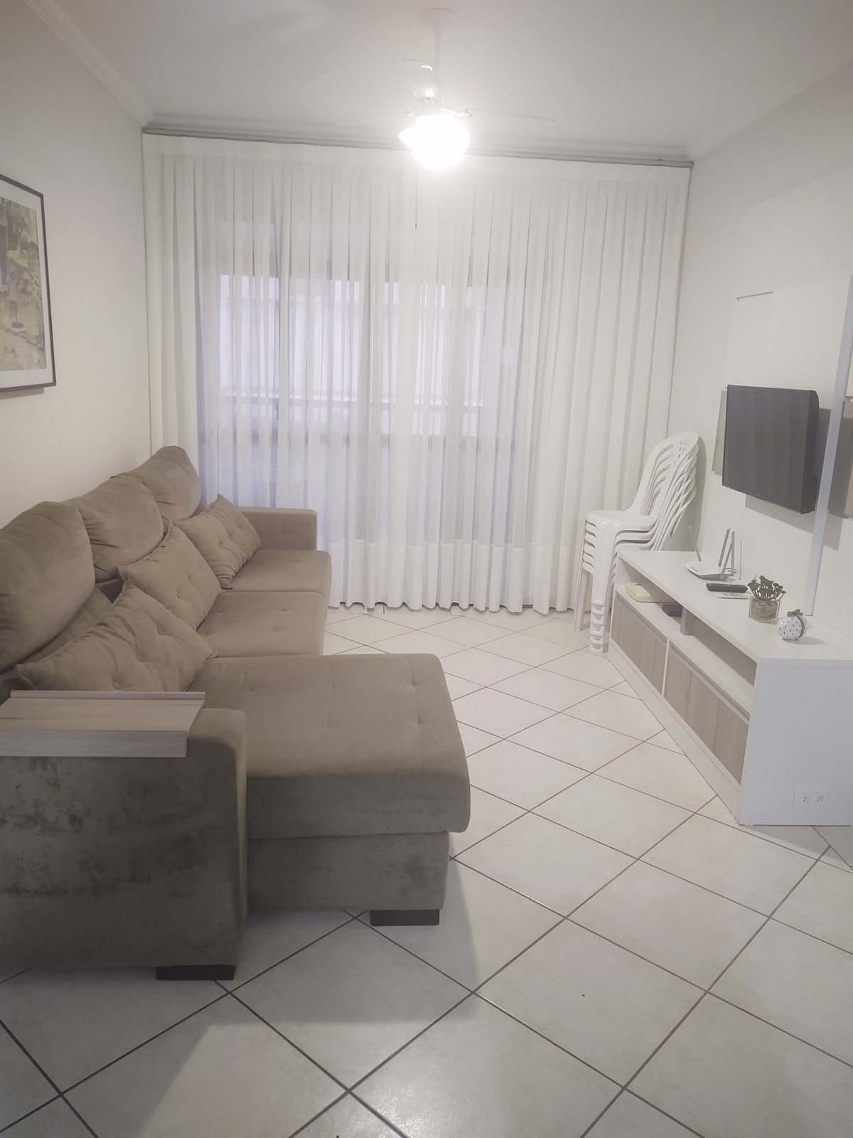 A light-filled living area features a plush sectional sofa in a neutral tone, complemented by a sleek entertainment unit. Large, sheer curtains frame a window, allowing natural light to filter in. A standing chair is placed nearby, contributing to the room's functional layout.