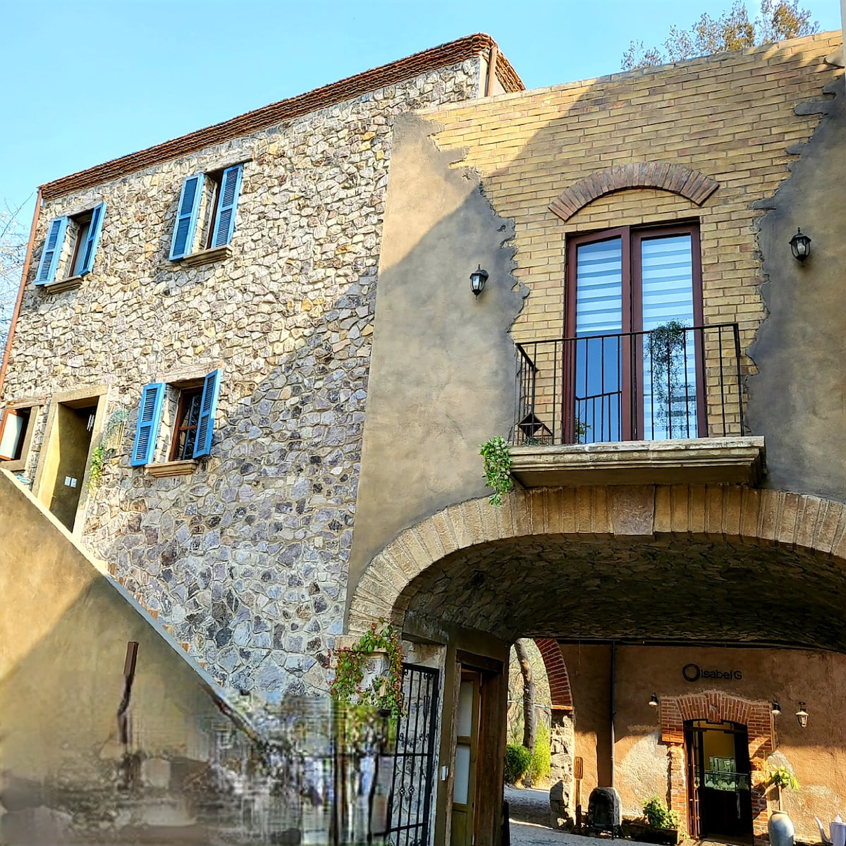 An elegantly structured building features a blend of stone and brick exteriors. Blue window shutters contrast with the natural stone. A curved archway provides access to a charming entrance, while a balcony above holds decorative plants, contributing to the architectural appeal.