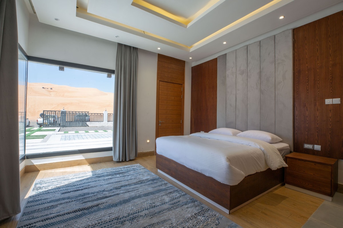 A spacious bedroom features a wooden bed positioned against a textured wall. Natural light floods the room through large glass doors, revealing a view of surrounding dunes. A plush area rug adds warmth to the wooden floor, enhancing the room's serene atmosphere.