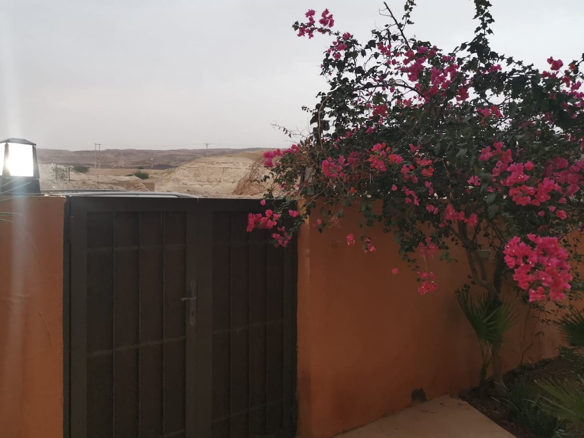 A decorative entrance features a wooden gate framed by vibrant flowering plants. The surrounding area showcases a natural landscape with distant hills, slightly obscured by a cloudy sky. A soft ambient light contributes to the serene atmosphere of the entrance.