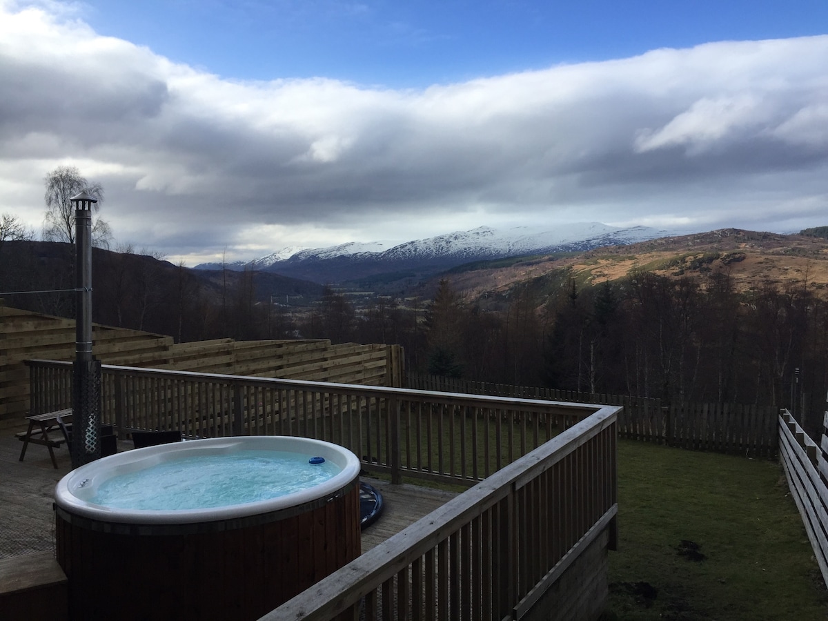 A wood-fired hot tub is positioned on a wooden deck, offering expansive views of the surrounding hills and valleys. Snow-capped mountains are visible in the distance, under a partly cloudy sky. The grassy area is enclosed by wooden fencing for added privacy.