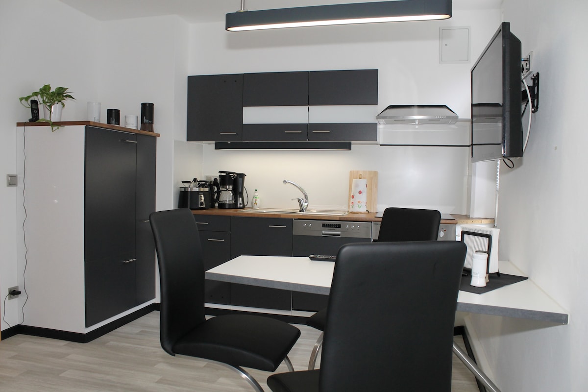 A modern kitchen is observed, featuring sleek dark cabinets and a wooden countertop. Equipped with essential appliances, including a coffee maker and dishwasher, a dining table with four black chairs is centrally placed, enhancing the kitchen's functional design.