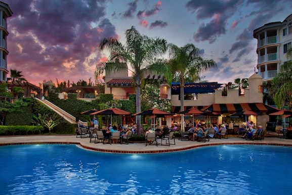 The resort's outdoor pool area is highlighted by inviting lounge seating shaded by large umbrellas. Guests can be seen enjoying refreshments, surrounded by lush green landscaping. The sky is adorned with hues of purple and orange at dusk, creating a relaxed atmosphere.