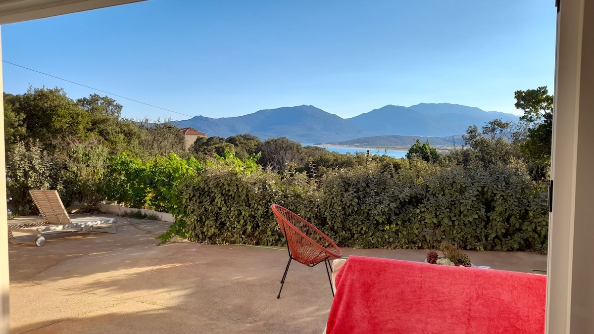 A spacious outdoor area features a vibrant red blanket draped over a seating arrangement. In the background, rolling hills and a serene body of water are visible. Natural greenery surrounds the space, enhancing the tranquil atmosphere.