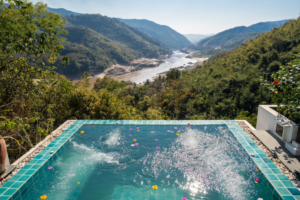 A tranquil Jacuzzi is positioned on a terrace, surrounded by lush greenery, offering a panoramic view of the Mekong River and distant mountains. Colorful flower petals float on the water's surface, enhancing the serene environment. The clear sky and natural landscape create a peaceful backdrop.