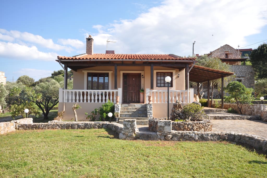 The outside of the house. It offers a spacious front yard with lawn, some olive trees and a parking space