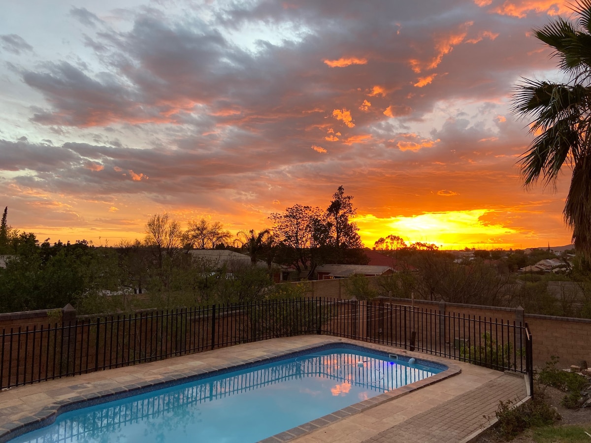 A serene outdoor scene captures a swimming pool framed by a black fence. The sky is illuminated by vibrant orange and pink hues during sunset, with dark clouds adding depth. Lush greenery is visible on the property, enhancing the natural setting.