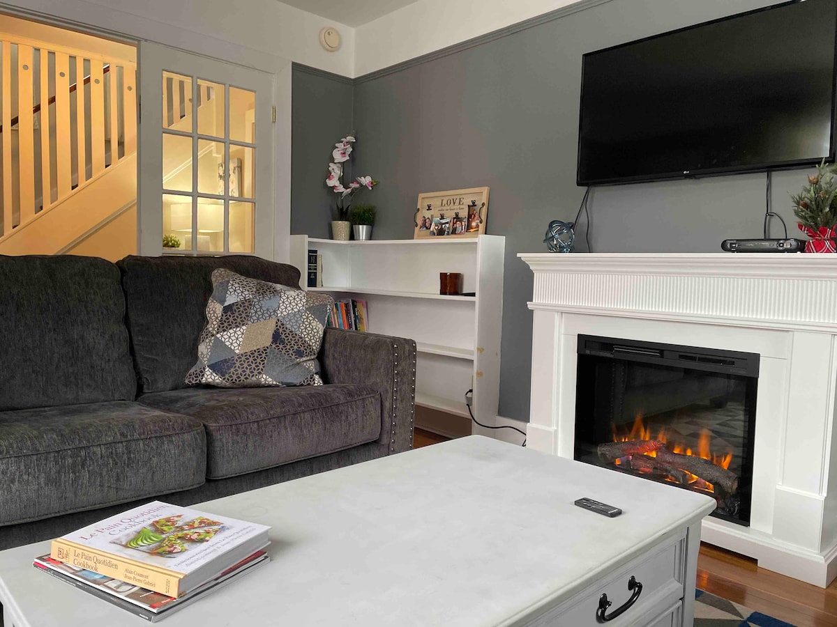 A cozy living room is depicted, featuring a dark gray sofa adorned with a patterned throw pillow. A white coffee table rests in front, displaying a magazine. A modern fireplace with an illuminated flame is situated beneath a mounted flat-screen TV. Light enters from a nearby staircase.