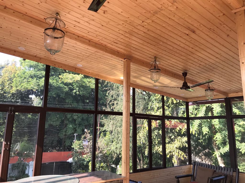 A sunlit terrace features large windows surrounded by wooden beams and a ceiling adorned with pendant lights. Lush greenery outside provides a vibrant backdrop, enhancing the open and airy atmosphere of the space.