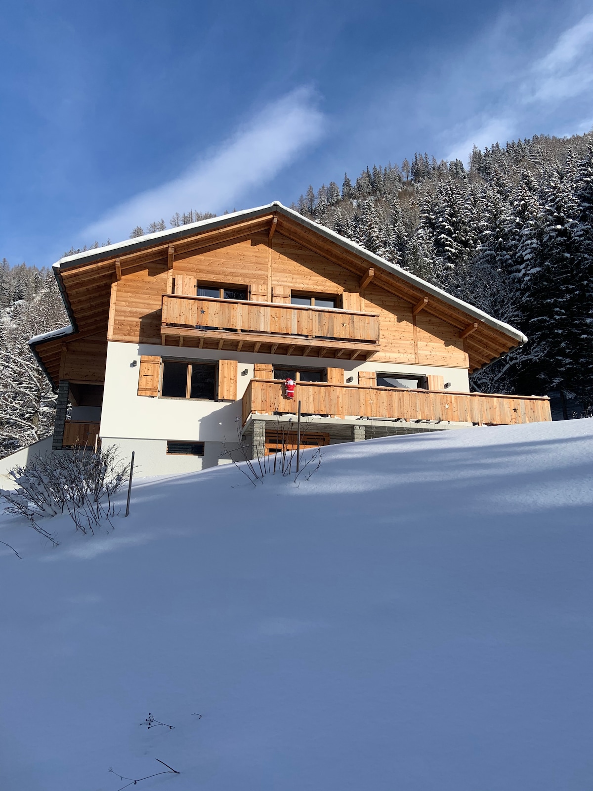 The chalet is nestled among tall trees, with a sloping roof and wooden balconies. The exterior showcases a blend of wood and white walls, surrounded by a blanket of snow. A clear blue sky provides contrast to the snowy landscape.