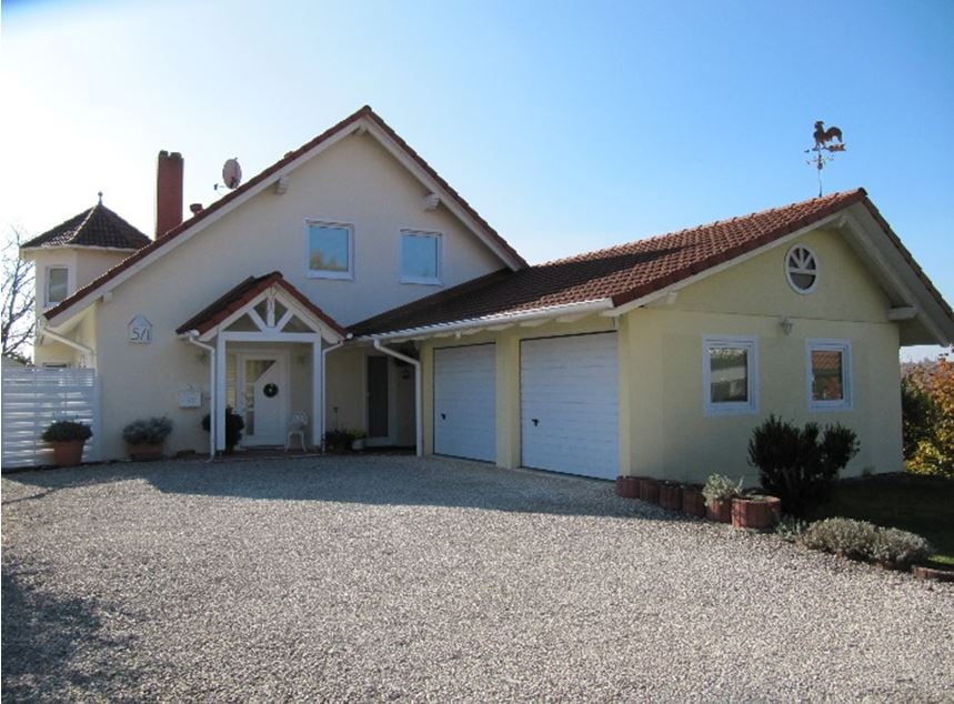 A two-story house displays a light-colored exterior with a pitched roof and decorative gables. A gravel driveway leads to two garages and a welcoming porch. Potted plants line the entrance, and a weather vane is positioned on the roof, capturing the essence of the rural surroundings.