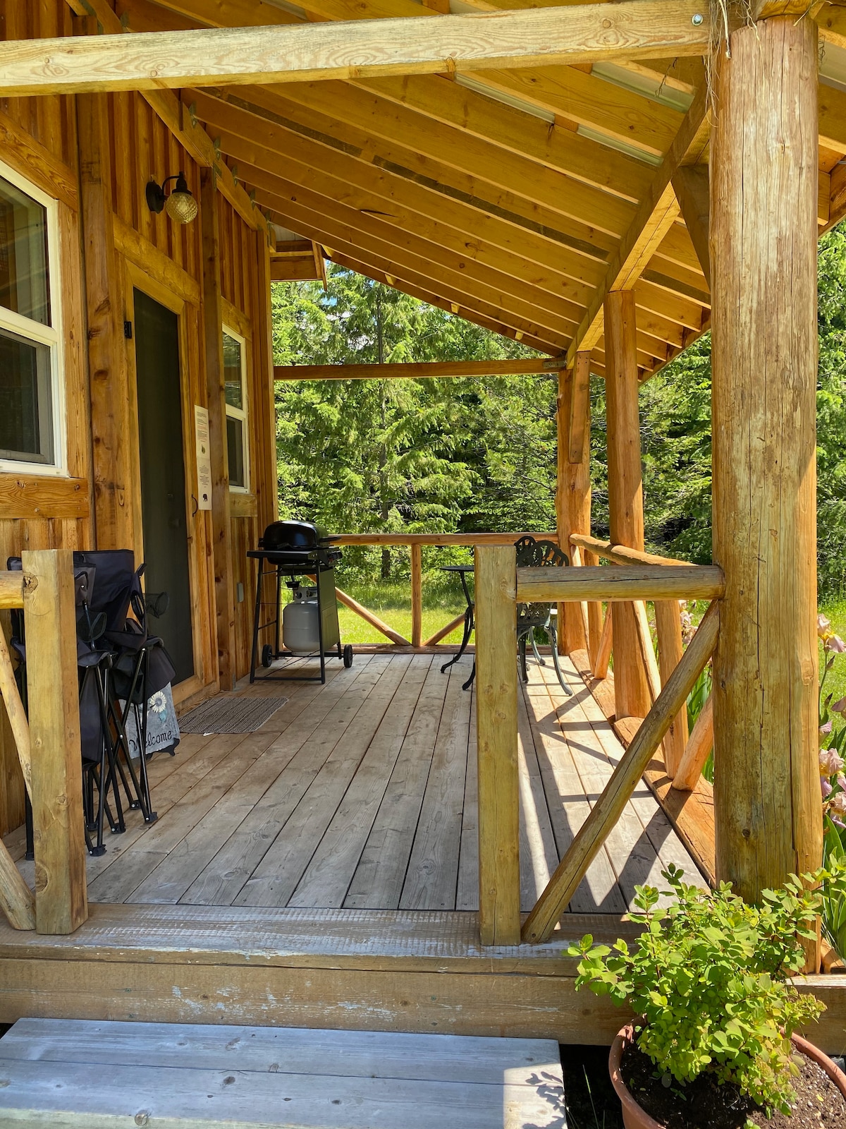 A wooden porch features a shaded area with seating chairs and a barbecue grill. The railings are made of rustic timber, and a sunny view of the surrounding greenery is visible beyond the porch. Potted plants add a touch of nature to the outdoor space.