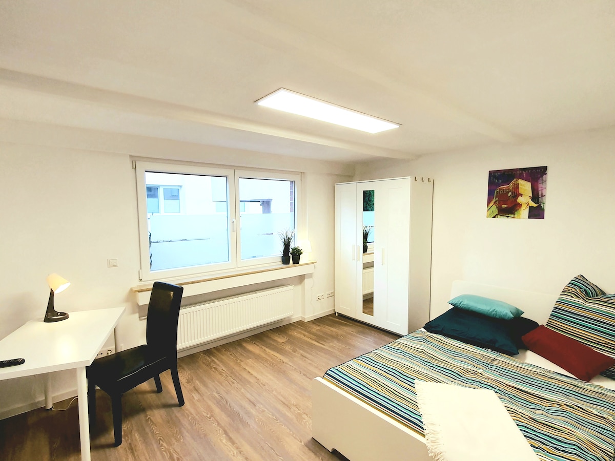 A spacious bedroom features a striped bedspread with coordinating pillows on a large bed. A desk with a lamp is positioned beside a bright window, providing natural light. A wardrobe and decorative wall art add to the functional design of the room.