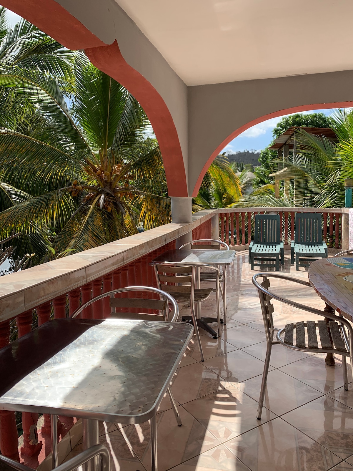 A spacious terrace overlooks lush palm trees, providing a relaxed outdoor setting. Metal tables and chairs are positioned for enjoyment of the surroundings. Sunlight reflects off the tiles, creating a bright atmosphere, while the archways frame the view, enhancing the sense of openness.