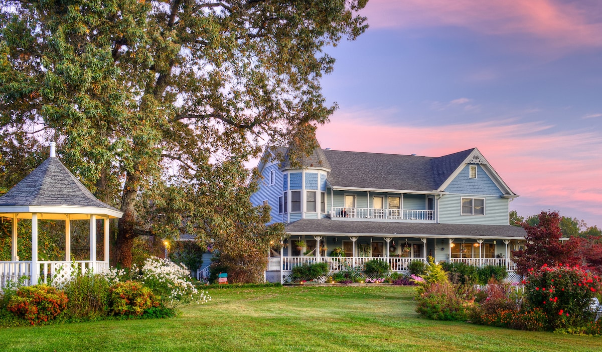 A charming blue house is set against a colorful sunset, featuring multiple balconies and a spacious porch. Surrounding gardens are filled with flowers and greenery. An adjoining gazebo is visible, complemented by a large tree providing shade in the foreground.
