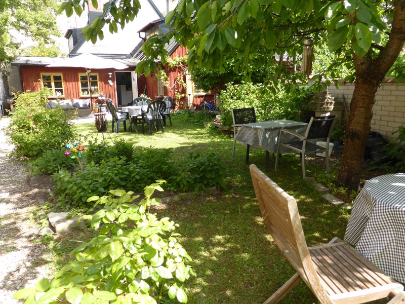 A lush garden space is presented, featuring several tables and chairs set on a grassy area. The vibrant greenery creates a serene environment, with a mix of paving stones and natural paths leading to a charming red building in the background.