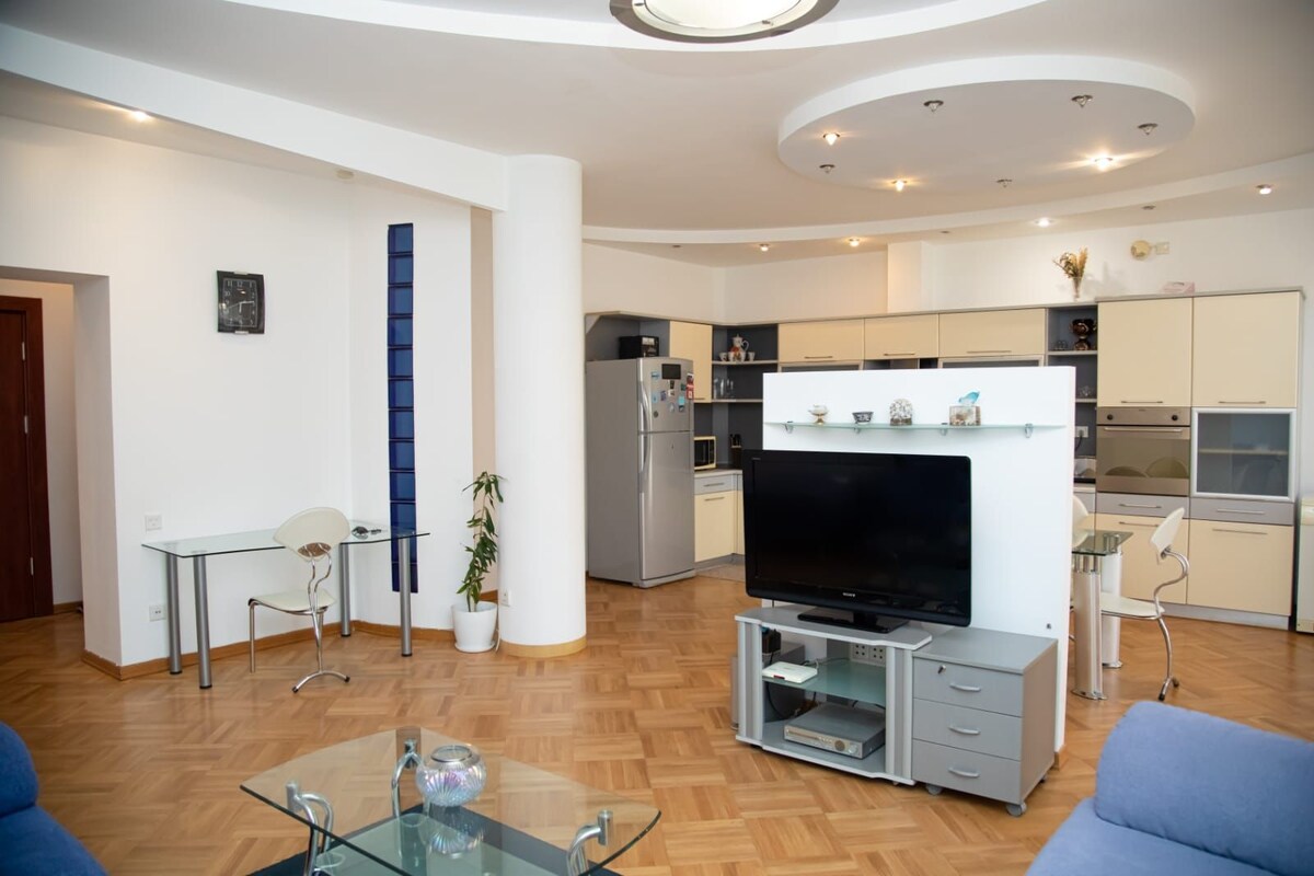 An open-concept living space features a modern kitchen with light cabinetry and a stainless steel refrigerator. A flat-screen TV is mounted on a display unit. A glass coffee table sits in the center, while a dining area with a table and chairs is visible in the background.