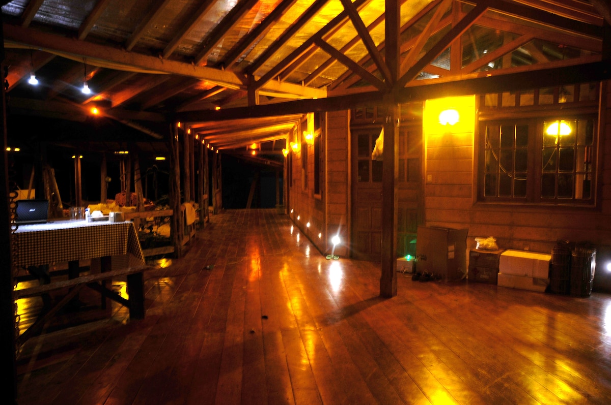 A spacious indoor area is illuminated with warm lighting, featuring polished wooden flooring and high wooden ceilings. Several dining tables are arranged along one side, while hints of furniture and decor create a welcoming atmosphere. The open layout provides flexibility for various activities.