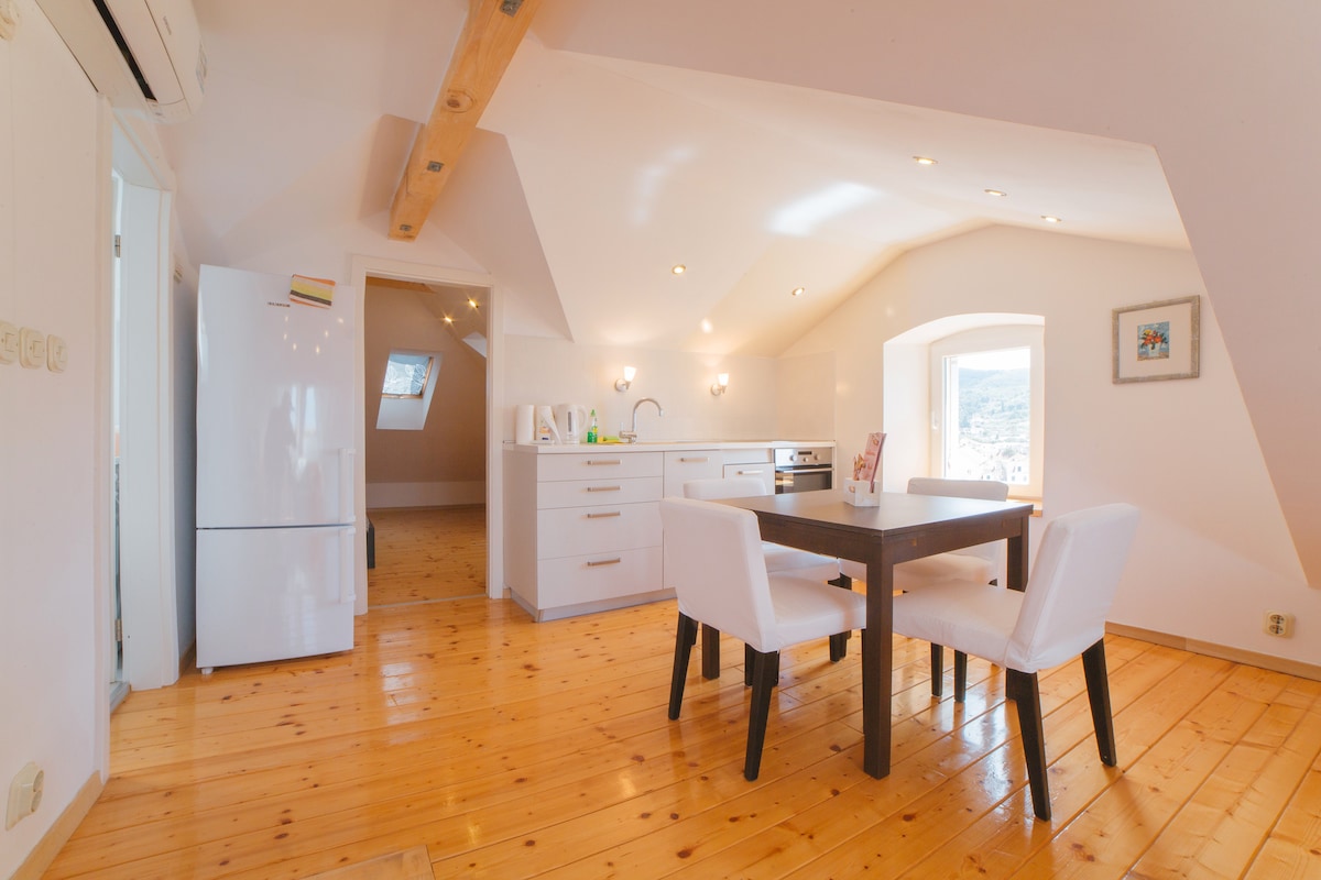 A well-lit dining area features a small table surrounded by four white chairs. A sleek kitchen is visible, equipped with modern appliances, including a refrigerator and a stove. Light wooden beams and a wooden floor contribute to a warm ambiance.