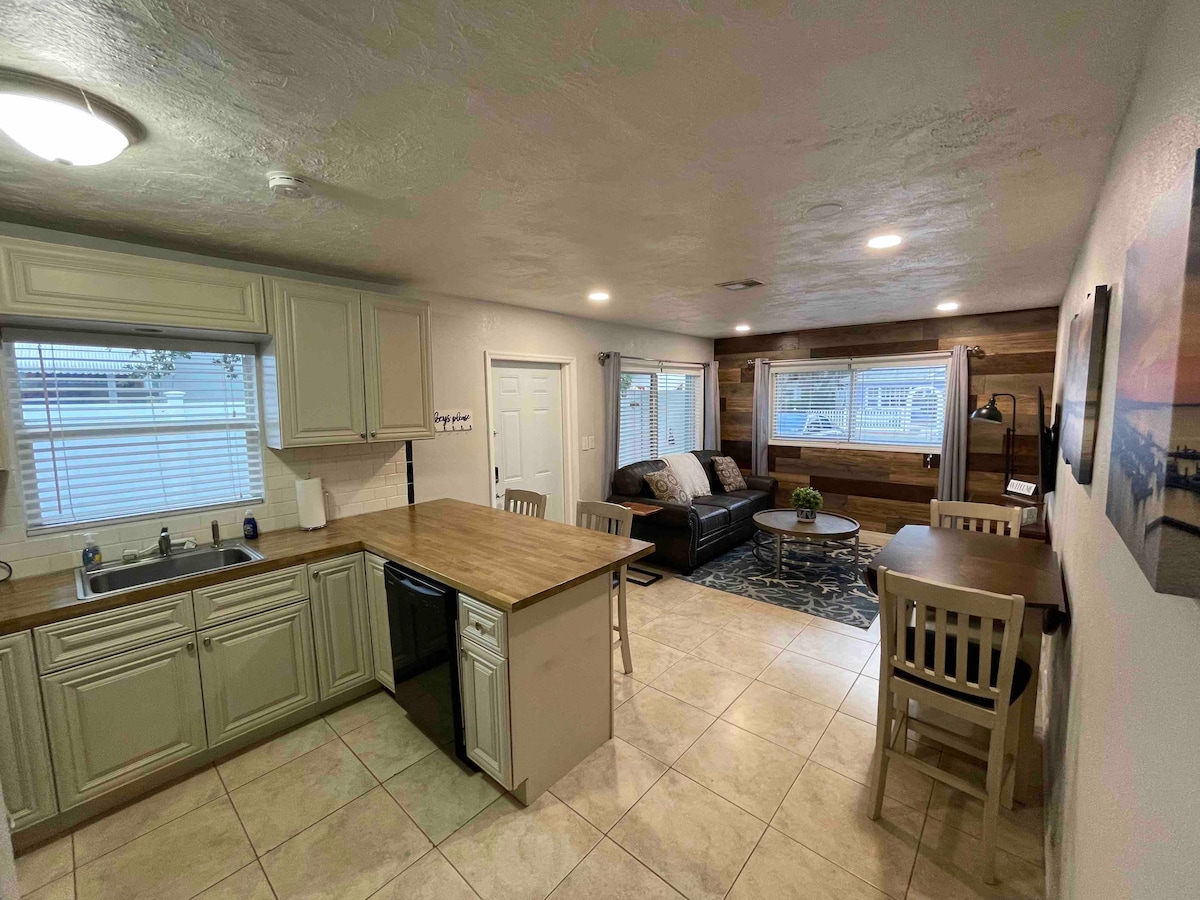 An open living area features a fully equipped kitchen with light-colored cabinets and a wooden countertop. A cozy seating area with a dark sofa is positioned alongside a round coffee table. Bright windows allow natural light to illuminate the space, while a small dining table accommodates two.