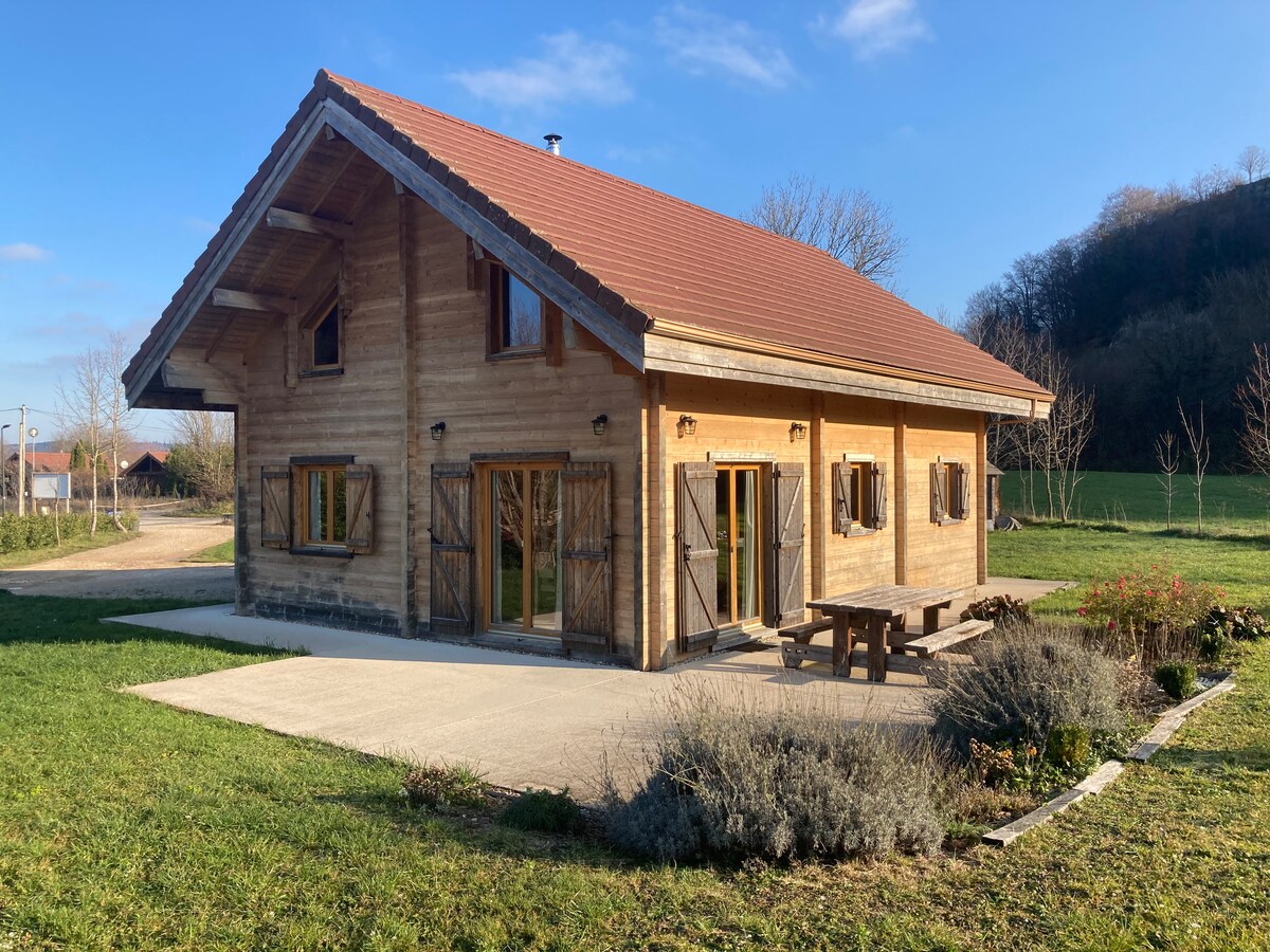 A spacious wooden chalet features a rustic design with wide eaves and large windows. A picnic table is situated on a concrete patio, surrounded by landscaped grass and shrubs. Nearby trees create a serene natural backdrop, complemented by a clear blue sky.