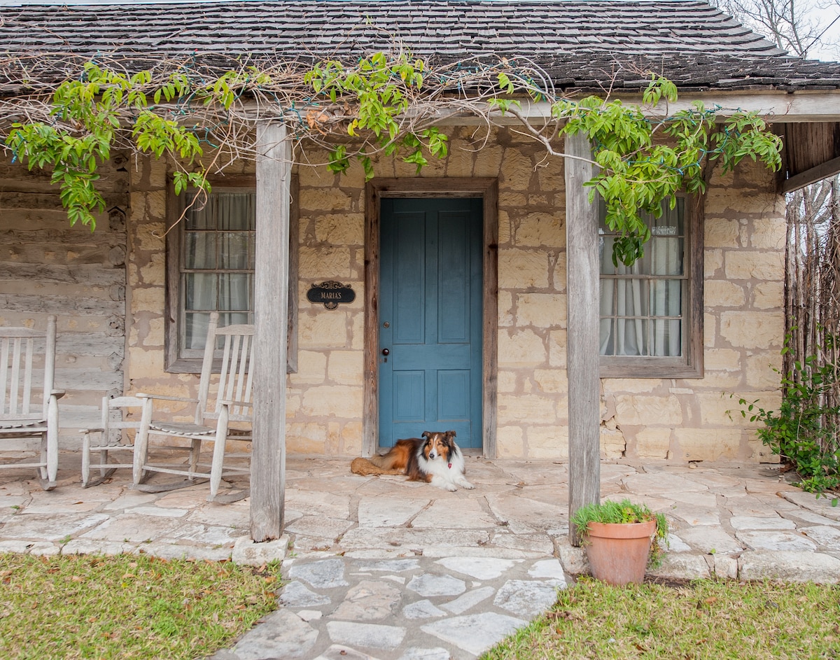 An inviting entrance to the log cabin features a blue front door framed by wooden pillars. Two rocking chairs are positioned on a stone patio, while a collie dog relaxes on the porch. Green climbing vines add a touch of nature to the rustic stone facade.