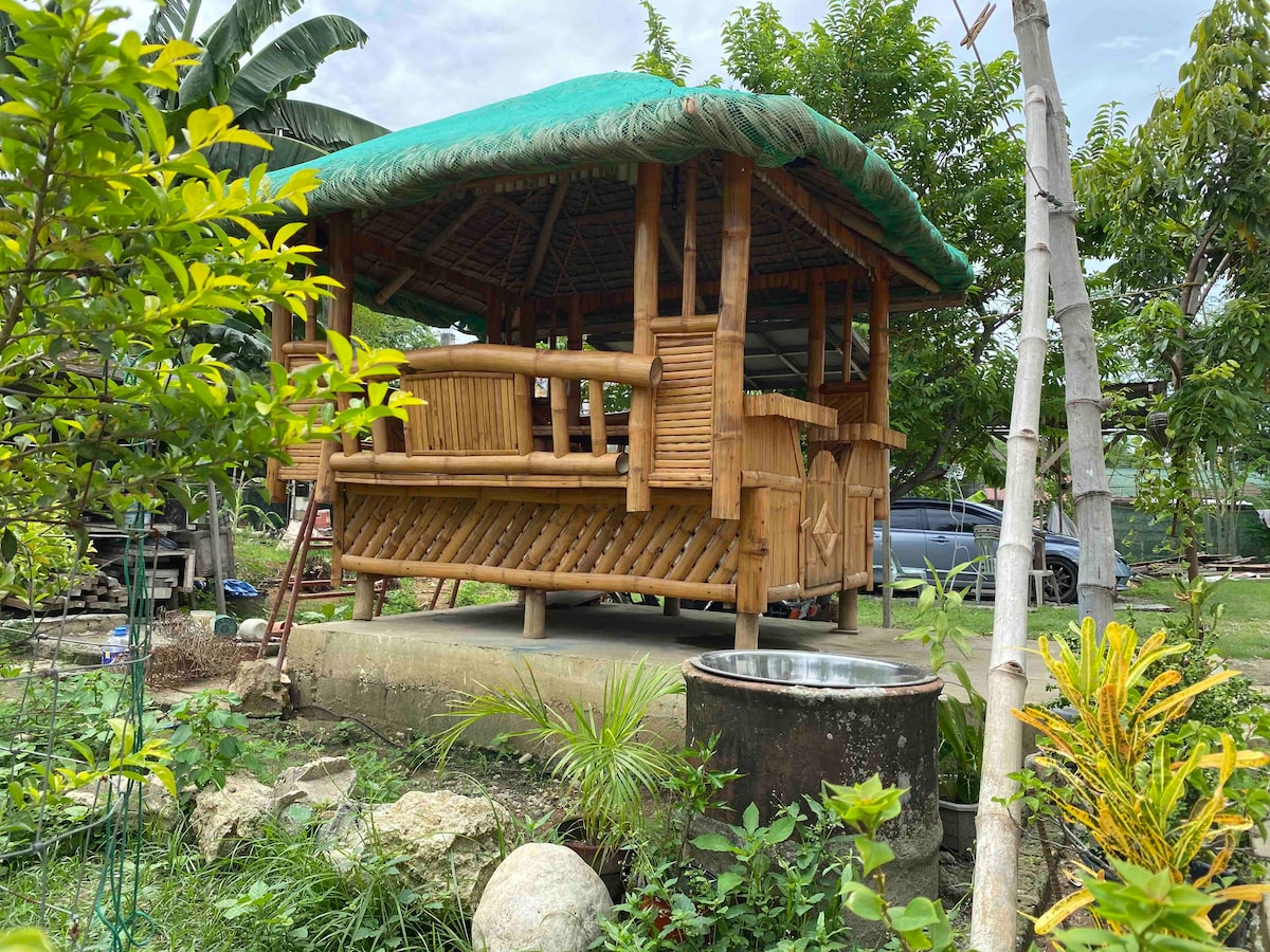 A traditional bamboo gazebo is positioned amid lush greenery. Its thatched roof provides shade, while the bamboo structure features benches for seating. Surrounding plants and stones create a natural environment, adding to the serene outdoor setting.