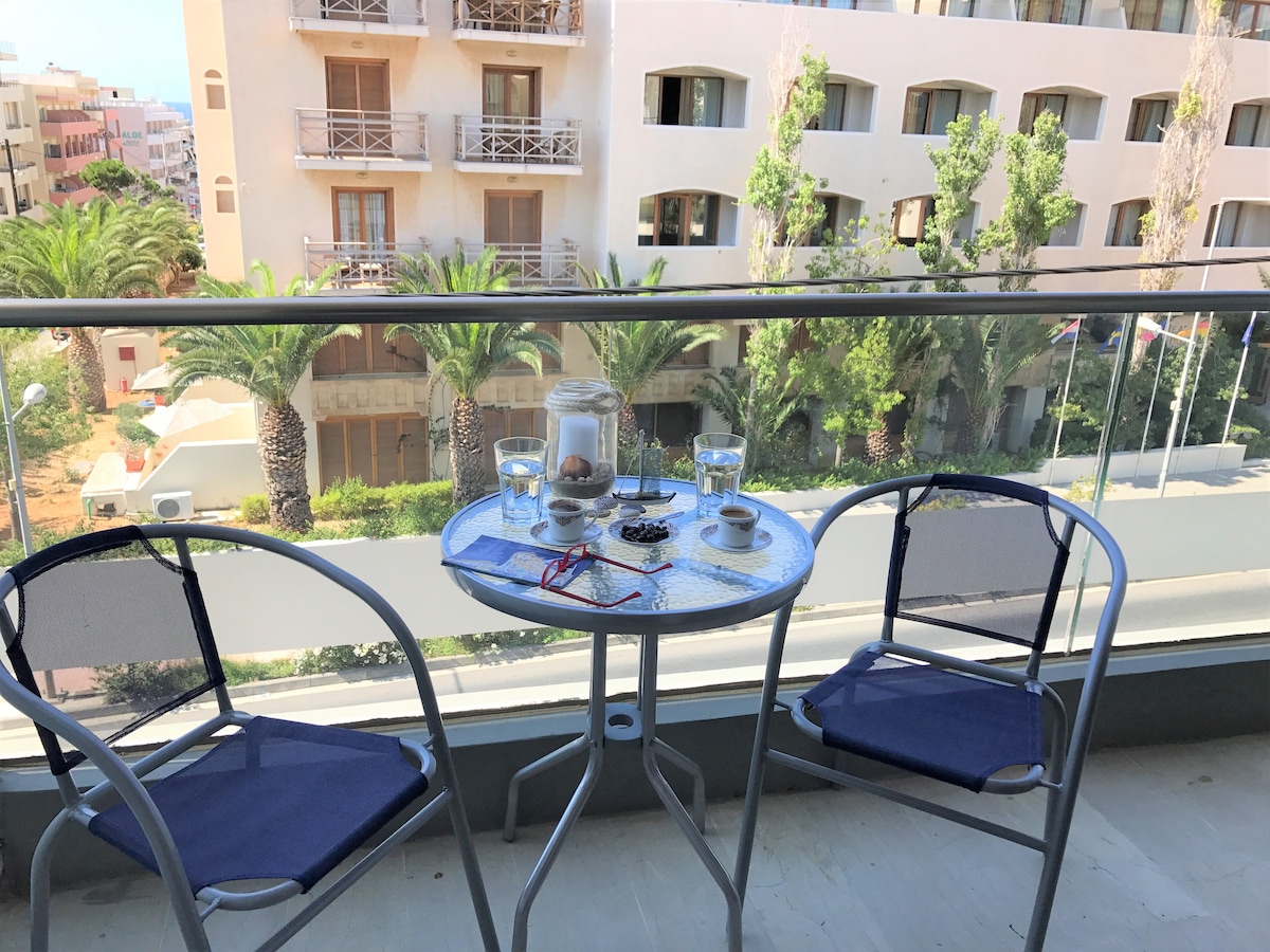 A small balcony is showcased with a round table and two chairs, providing a space to relax. Glasses and an arrangement of items sit on the table. Lush palm trees and buildings can be seen in the background, adding to the scenic view.