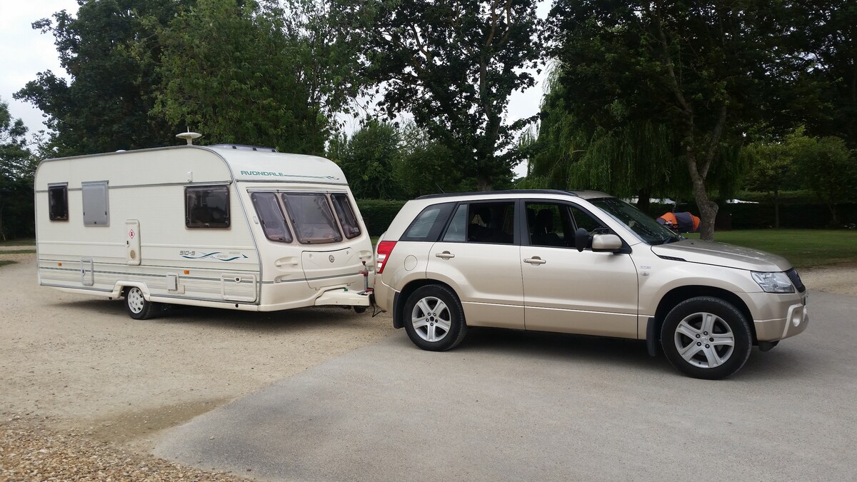 A light-colored caravan is parked alongside a beige SUV in a grassy area surrounded by trees. The caravan features simple lines and a compact design, while the SUV showcases five-spoke wheels and a sleek profile. Natural surroundings provide a peaceful backdrop.