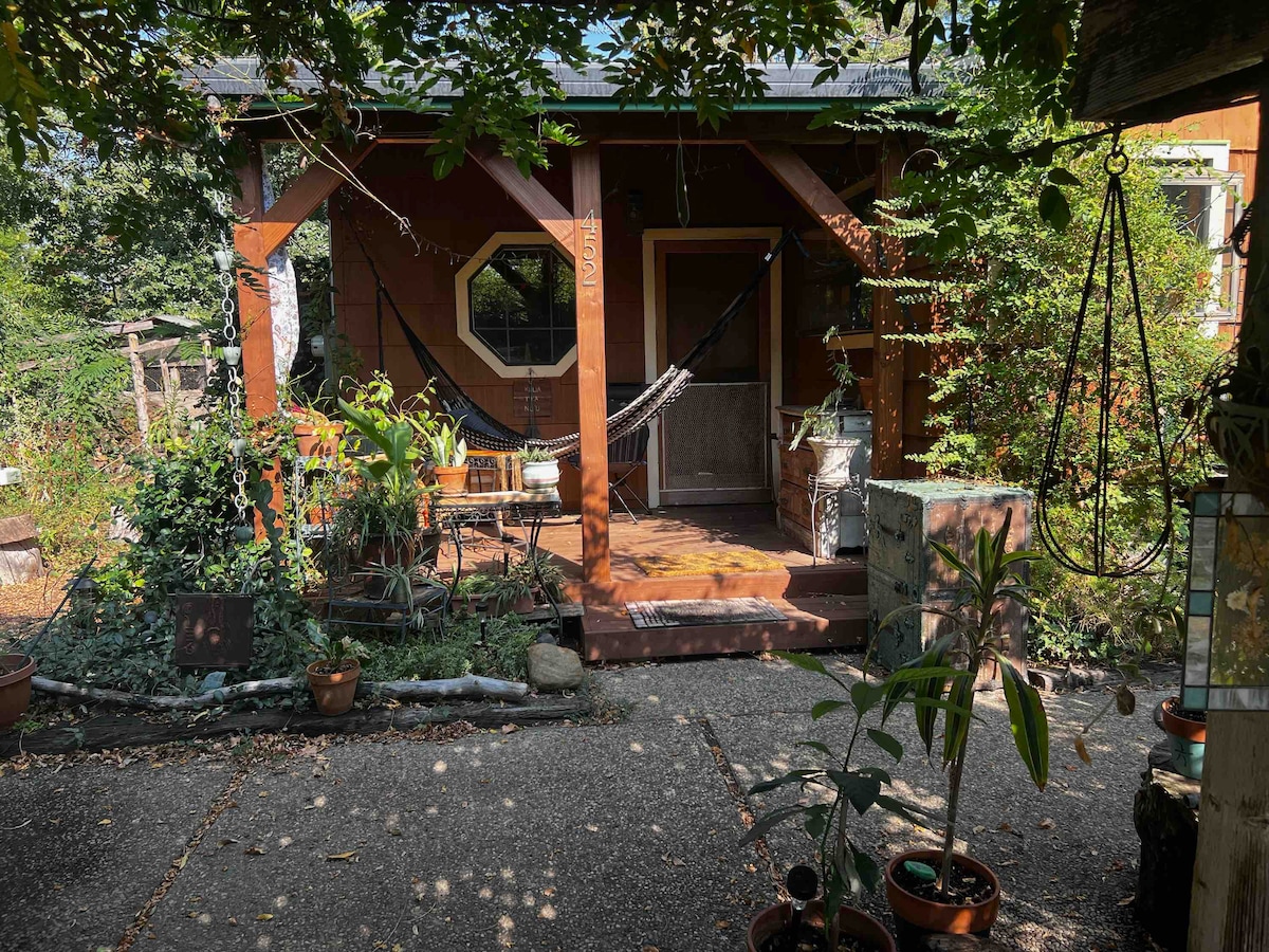 A cozy porch area is depicted, featuring a hammock suspended between wooden posts. Potted plants are arranged along the path, complementing the green surroundings. The entrance is framed by foliage, creating a welcoming atmosphere, while a rustic wooden structure is visible in the background.