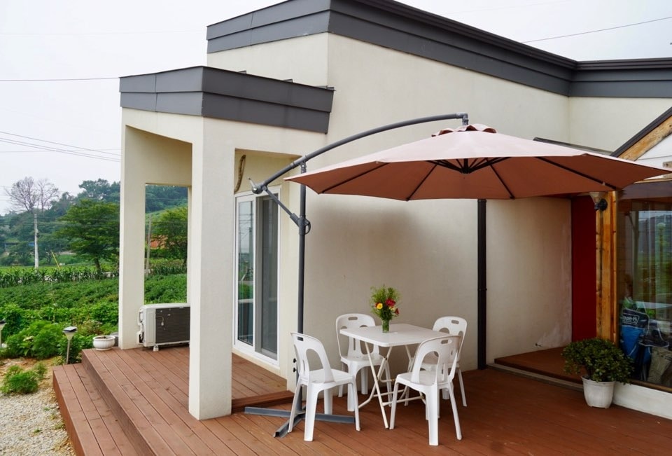 An outdoor seating area features a round table surrounded by four white chairs, shaded by a large umbrella. Potted plants add greenery, while a wooden deck extends around the area, providing a peaceful space for relaxation.