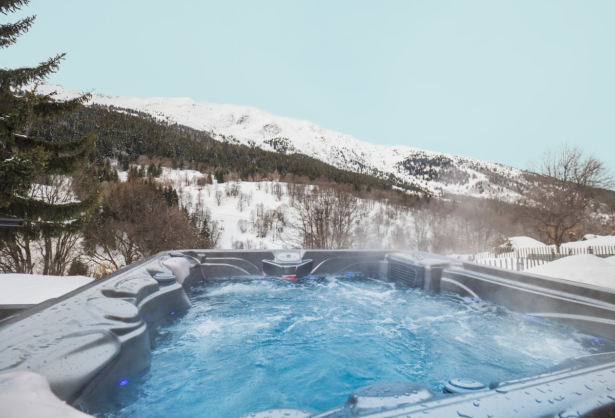 A hot tub filled with steaming water is set against a backdrop of snow-covered mountains. The clear sky above offers a serene atmosphere. The surrounding landscape is dotted with trees, enhancing the peaceful outdoor setting.