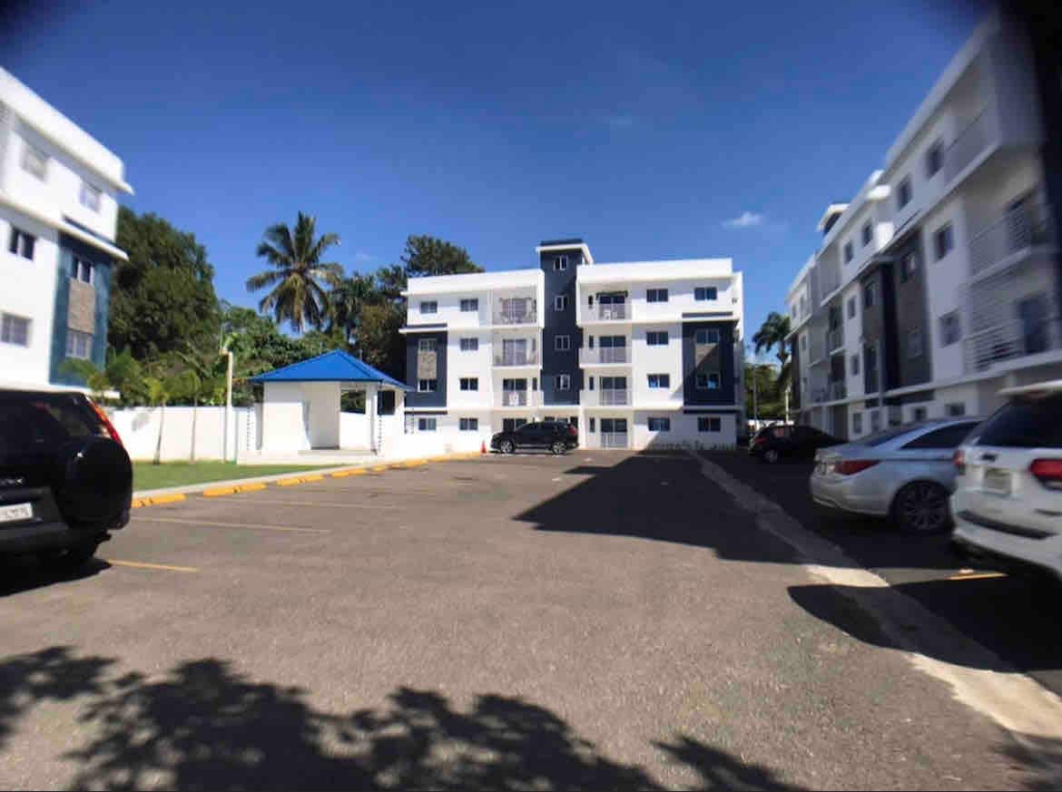 A gated apartment complex is depicted with several three-story buildings featuring blue and white exteriors. A designated parking area is visible, accommodating multiple vehicles. A gazebo is positioned in the background, surrounded by greenery, providing an inviting outdoor space.